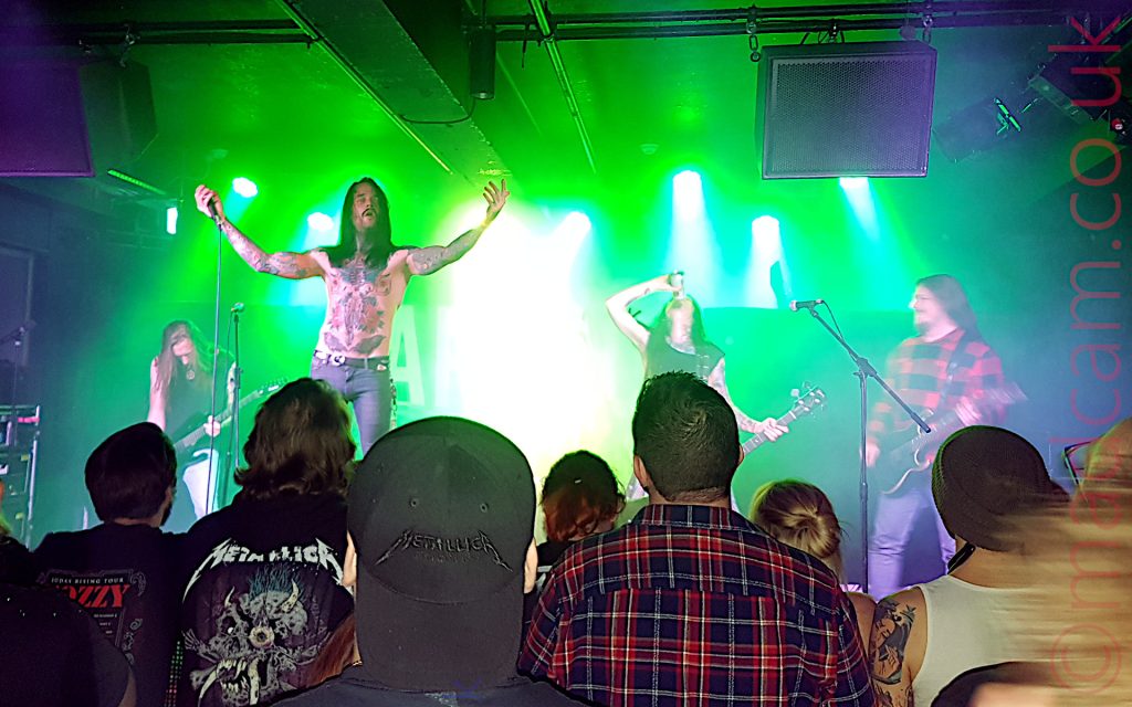 4 members of a heavy metal band on stage. On the left, at the back, is a man with long blond hair wearing a dark shirt and playing a black electric guitar, his head bent downwards. In the middle, at the front of the stage, is a man with long dark hair with a moustache and the beginnings of a beard, his bare chest covered with tattoos, and wearing a dirty pair of blue jean, holding a microphone in his right hand, his arms stretched out to the side in supplication. In the middle, towards the back of the stage, is a man with long dark hair, wearing a sleeveless black shirt, and holding the neck of a bass guitar in his left hand, his right hand holding a can, pouring it's contents down his raised throat. On the right is a man with long dark hair and a wispy beard, wearing a red and black checked jacket, playing a black electric guitar and staring across to the left of the frame. The left and right sides of the stage are lit blue by the stage lights, with most of it flooded with green light, and heavily over-exposed white lights mostly pointing in the general direction of down. In the foreground, the backs of the audiences heads can be seen, various band hoodies or checked shirts being common forms of attire.