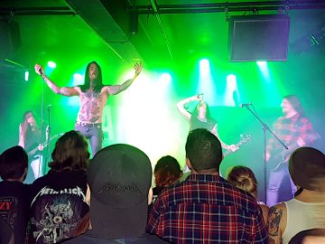 4 members of a heavy metal band on stage. On the left, at the back, is a man with long blond hair wearing a dark shirt and playing a black electric guitar, his head bent downwards. In the middle, at the front of the stage, is a man with long dark hair with a moustache and the beginnings of a beard, his bare chest covered with tattoos, and wearing a dirty pair of blue jean, holding a microphone in his right hand, his arms stretched out to the side in supplication. In the middle, towards the back of the stage, is a man with long dark hair, wearing a sleeveless black shirt, and holding the neck of a bass guitar in his left hand, his right hand holding a can, pouring it's contents down his raised throat. On the right is a man with long dark hair and a wispy beard, wearing a red and black checked jacket, playing a black electric guitar and staring across to the left of the frame. The left and right sides of the stage are lit blue by the stage lights, with most of it flooded with green light, and heavily over-exposed white lights mostly pointing in the general direction of down. In the foreground, the backs of the audiences heads can be seen, various band hoodies or checked shirts being common forms of attire.