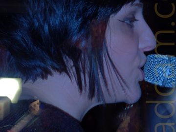 Close-up of the head of a woman facing to the right, singing in to a microphone on a stand right in front of her mouth. She has short, dark hair cut in a bob, and is wearing a dark top. Stage lights give her face a purple cast. A long exposure with a snap zoom results in this closeup of her face, but also a shot of her playing a creamy yellow guitar, with someone behind her playing a bass guitar in front of a bright light.