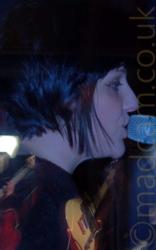 Close-up of the head of a woman facing to the right, singing in to a microphone on a stand right in front of her mouth. She has short, dark hair cut in a bob, and is wearing a dark top. Stage lights give her face a purple cast. A long exposure with a snap zoom results in this closeup of her face, but also a shot of her playing a creamy yellow guitar, with someone behind her playing a bass guitar in front of a bright light.