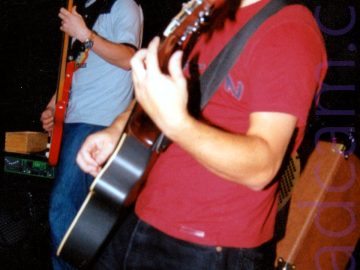 A view from the side of a man with short, curly dark hair, wearing a red t-shirt and a black pair of jeans, and playing a black acoustic guitar, looking down at his left hand as he stands facing off to the left of the frame. An orange speaker is mounted at an angle behind him, while a taller man with short dark hair and wearing a light and dark blue t-shirt is playing a red bass guitar off to the left of the frame. Black walls and ceiling fill the rest of the frame.