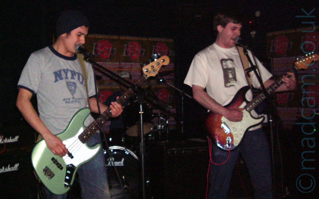 Two men performing on stage as part of a rock band. On the left is a man wearing a black knitted hat over short dark hair, with a grey t-shirt with black piping and an "NYPD" logo, and playing a green and white bass guitar. On the right is a man with short dark hair, wearing a white t-shirt with an indistinct black logo, and playing a brown and white electric guitar. Both are singing into microphones mounted on stands in front of them. Behind them, in the middle of he frame, is a drumkit, with the drummer barely visible, apart from a blue and white truckers cap. Brown posters with a red seal and with grey text "The Bop" cover most of the black walls in the background.