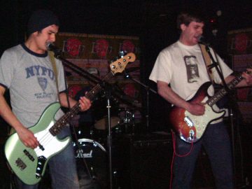 Two men performing on stage as part of a rock band. On the left is a man wearing a black knitted hat over short dark hair, with a grey t-shirt with black piping and an "NYPD" logo, and playing a green and white bass guitar. On the right is a man with short dark hair, wearing a white t-shirt with an indistinct black logo, and playing a brown and white electric guitar. Both are singing into microphones mounted on stands in front of them. Behind them, in the middle of he frame, is a drumkit, with the drummer barely visible, apart from a blue and white truckers cap. Brown posters with a red seal and with grey text "The Bop" cover most of the black walls in the background.