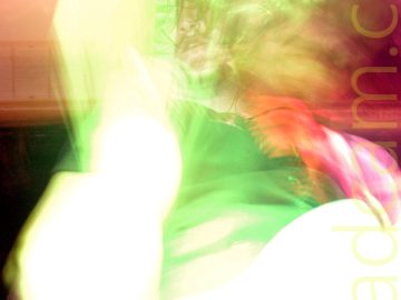 Long exposure shot of a man with long, shaggy, dark hair, wearing a black t-shirt and a loosely knotted red and black striped tie, and playing a white bass guitar. The long exposure, his movement, and the stage lights combine to leave his face perfectly focused but leaves everything else either blown out (his bass and right arm) or smeared to the point of almost being un-recognisable (his bouncing hair, left shoulder, the background). An explosion of white light has left just a vague shape where his bass and right arm should be, while green lights highlight the rest of him, and red lights highlight parts of the black wall in the background.