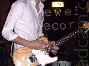 A man with dark, curly hair, wearing a white shirt and faded blue jeans, and playing a brown and white electric guitar, while facing the right of the frame. A white drumkit is on the right of the frame behind him, under a black sign on the wall, with only the letters "EWEI" and "ECOR" visible, under a bright yellowish light. Black walls and ceiling fill the rest of the frame.