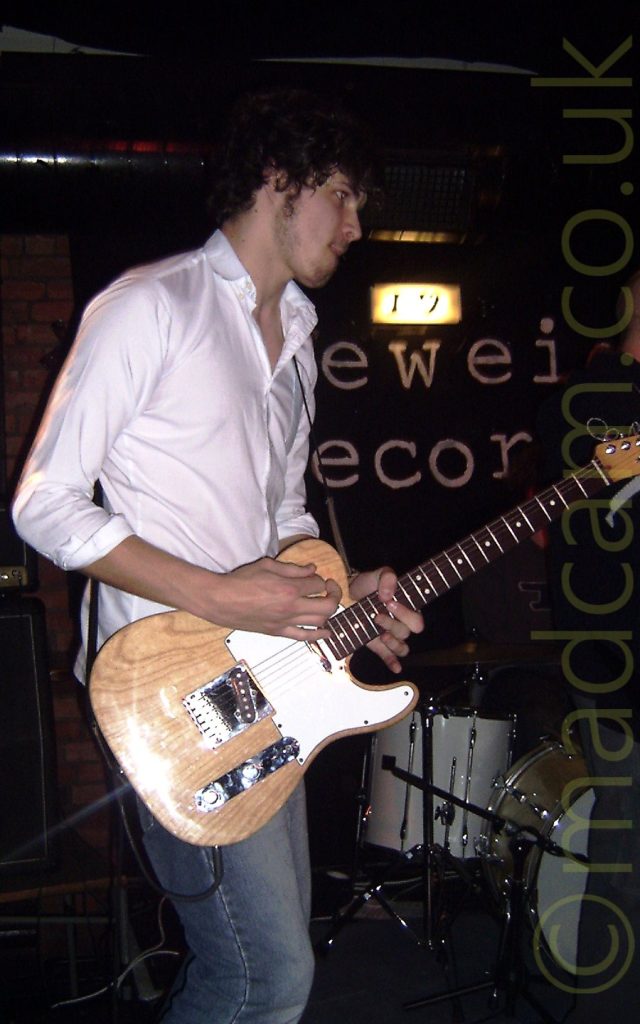 A man with dark, curly hair, wearing a white shirt and faded blue jeans, and playing a brown and white electric guitar, while facing the right of the frame. A white drumkit is on the right of the frame behind him, under a black sign on the wall, with only the letters "EWEI" and "ECOR" visible, under a bright yellowish light. Black walls and ceiling fill the rest of the frame.