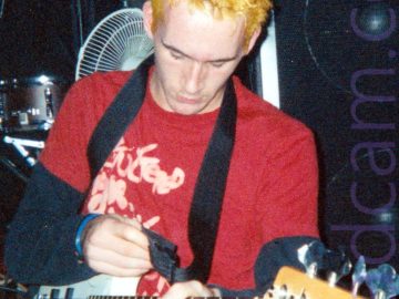 A man with spiky yellow hair, wearing a red t-shirt with indistinct white text over a black long-sleeved t-shirt, and a dark pair of trousers, on his knees on the floor, his feet splayed out behind him. He is playing a white bass guitar that is attached to a black strap over his left shoulder. A large black Peavey amplifier fills the background on the right, with part of a drumkit and a white desktop fan are against the black wall on the left.