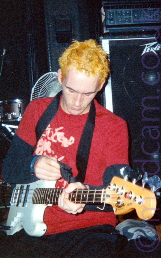 A man with spiky yellow hair, wearing a red t-shirt with indistinct white text over a black long-sleeved t-shirt, and a dark pair of trousers, on his knees on the floor, his feet splayed out behind him. He is playing a white bass guitar that is attached to a black strap over his left shoulder. A large black Peavey amplifier fills the background on the right, with part of a drumkit and a white desktop fan are against the black wall on the left.