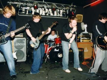 Five members of a rock band performing on stage. On the left is a man with mid-length blond hair, wearing a dark blue t-shirt and blue jeans, and playing a black electric guitar. Next to him is a man with dark hair, wearing a black t-shirt and baggy blue jeans, playing a black bass guitar. In the middle, at the back of the stage, is a man with short dark hair, wearing a white t-shirt, and sitting behind a red drum kit, the stick in his left hand slightly raised in to the air. Next is a man with dark hair, wearing a black t-shirt and the almost obligatory blue jeans, slightly hunched over as he sings into a microphone he is holding in his right hand. On the right is a man with dark hair, wearing a long-sleeved black top with a white stripe down the arm, and - Shock! Horror! - a grey pair of trousers with a black stripe down the side of the leg., and playing a black electric guitar. Various amplifiers and speakers (and a white electrical fan aimed at the drummer) litter the back of the stage. Black walls and ceiling fill the background, with large white text "The Leopard" high up on the wall at the back, under a metal lighting rig.