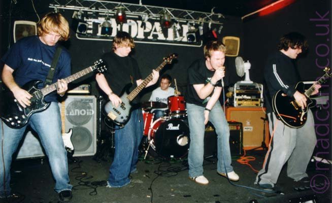 Five members of a rock band performing on stage. On the left is a man with mid-length blond hair, wearing a dark blue t-shirt and blue jeans, and playing a black electric guitar. Next to him is a man with dark hair, wearing a black t-shirt and baggy blue jeans, playing a black bass guitar. In the middle, at the back of the stage, is a man with short dark hair, wearing a white t-shirt, and sitting behind a red drum kit, the stick in his left hand slightly raised in to the air. Next is a man with dark hair, wearing a black t-shirt and the almost obligatory blue jeans, slightly hunched over as he sings into a microphone he is holding in his right hand. On the right is a man with dark hair, wearing a long-sleeved black top with a white stripe down the arm, and - Shock! Horror! - a grey pair of trousers with a black stripe down the side of the leg., and playing a black electric guitar. Various amplifiers and speakers (and a white electrical fan aimed at the drummer) litter the back of the stage. Black walls and ceiling fill the background, with large white text "The Leopard" high up on the wall at the back, under a metal lighting rig.