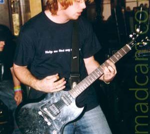 A man with floppy blond hair, wearing a black t-shirt with the white text "Help me find my", the last word of this phrase hidden behind his black guitar strap, slung over his shoulder. A blue pair of jeans and a black electric guitar complete the ensemble. Wooden cabinets with glass-fronted doors mounted on the wall fill the background.