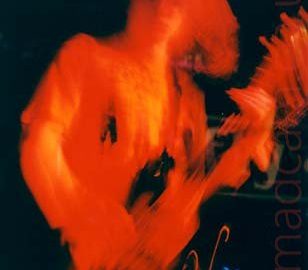 A man with floppy dark hair, playing a bass guitar on stage. The stage lighting has painted him entirely red, which combined with a long exposure and his movement has left red trails, particulary his hair and the head of his bass. Parts of the drum kit are illuminated behind him, with black walls filling the rest of the frame.