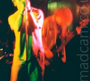 A man with short dark hair, wearing a light-coloured t-shirt and grey shorts, leaning on a microphone stand as he holds the mic in both hands as he sings into it. Stage lights paint his right side yellow, and his left side red and pink, with green in the middle. Behind him, on the right, is another man with shaggy hair, wearing an open grey shirt and playing a black bass guitar, slightly hunched over as he plays. Black walls, some flooded with green of red lights, and black ceiling, fill the background.