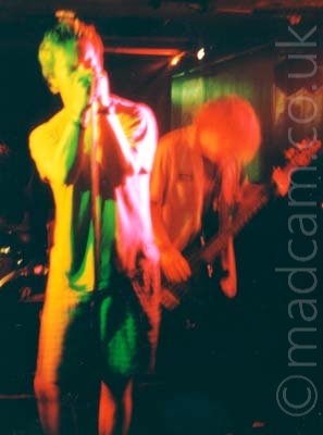 A man with short dark hair, wearing a light-coloured t-shirt and grey shorts, leaning on a microphone stand as he holds the mic in both hands as he sings into it. Stage lights paint his right side yellow, and his left side red and pink, with green in the middle. Behind him, on the right, is another man with shaggy hair, wearing an open grey shirt and playing a black bass guitar, slightly hunched over as he plays. Black walls, some flooded with green of red lights, and black ceiling, fill the background.