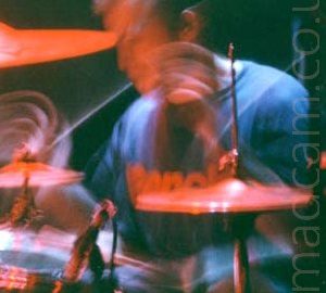 A man with short dark hair, wearing a dark blue t-shirt with indistinct red text across the chest, sitting behind a black and chrome drum kit. A long exposure and his movement has streaks of light as his drumsticks move, lit red by the stagelights, along with the rest of the kit and his face. Black walls fill the background.