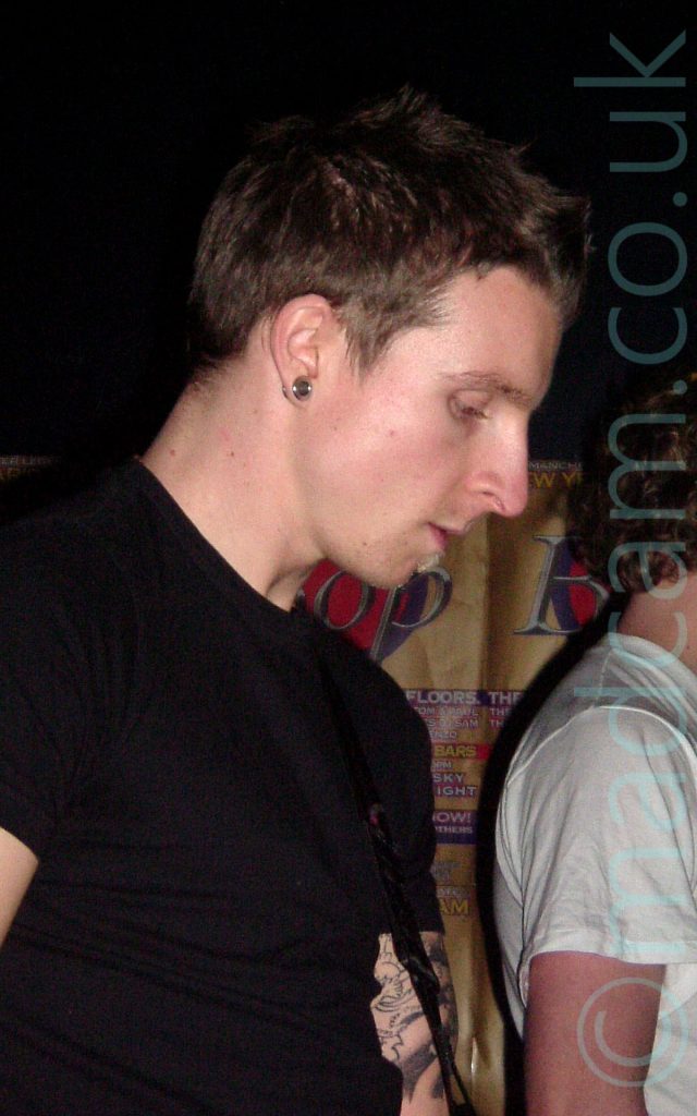 Close-up of the head and shoulders of a man with short, spiky brown hair, wearing a black t-shirt, with a black guitar strap over his left shoulder. The shoulders and back of the head of another man is on the right of the frame in the background. Gold-coloured posters for an event called "Bop" adorn the black walls behind them.