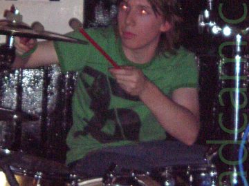 A man with floppy brown hair, wearing a green t-shirt and blue jeans, sitting behind a gold-coloured drum kit, holding a couple of red drums sticks up to play a metal cymbal. A large white poster on the black wall has a large, black, stylised letter "W" in the bottom right corner.