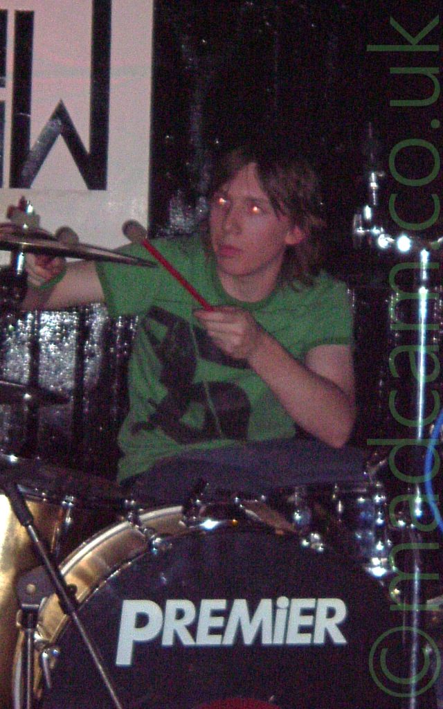 A man with floppy brown hair, wearing a green t-shirt and blue jeans, sitting behind a gold-coloured drum kit, holding a couple of red drums sticks up to play a metal cymbal. A large white poster on the black wall has a large, black, stylised letter "W" in the bottom right corner.