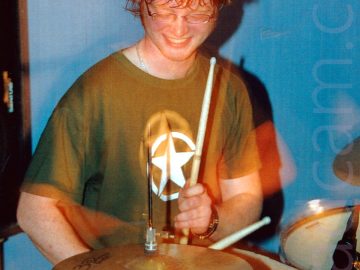 A man with lank brown hair and a pair of eye-glasses, wearing a khaki green shirt with a white star in the white outline of a circle, and a blue pair of jeans. He is sitting behind a black and chrome drum kit, with a drumstick lightly raised in the air, about to hit a cymbal. A long exposure has red secondary images where his head and arms have moved. Blue walls fill the background.