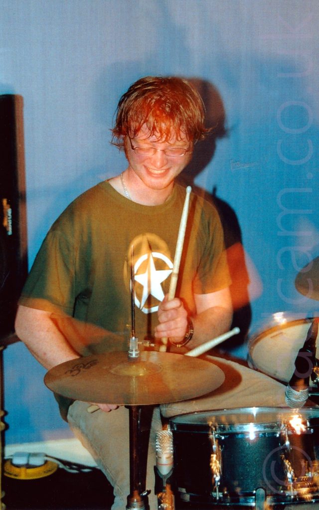 A man with lank brown hair and a pair of eye-glasses, wearing a khaki green shirt with a white star in the white outline of a circle, and a blue pair of jeans. He is sitting behind a black and chrome drum kit, with a drumstick lightly raised in the air, about to hit a cymbal. A long exposure has red secondary images where his head and arms have moved. Blue walls fill the background.