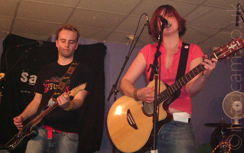 Two people performing on a stage as part of a band. On the left is a man with short brown hair, wearing a black t-shirt with a yellow and red text logo and a blue pair of jeans, playing a black electric guitar attached to a black and brown strap slung over his left shoulder. On the right is a woman with hair cut in a red bob, wearing a pink top and a pair of faded, dark blue jeans, playing a golden-brown acoustic guitar attached to a black strap over her left shoulder She is singing into a microphone on a stand in front of her. A black cloth with the letters "Sa" at the start of a word in white, the rest hidden behind Greg, is on the purple wall behind them, with a tiled white ceiling at the top of the frame.