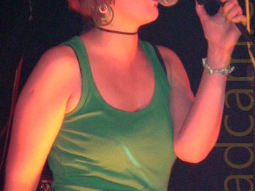 A woman with short, red hair cut in a bob, wearing a green tank top and blue jeans, singing into a microphone on a stand in front of her. She is holding the microphone in her left hand, with her right hand down by her leg. Her eyes are shut, showing that she is wearing makeup on her upper eye-lid. She also has green, circular, dangly ear-rings, and a thin black choker around her neck. The lighting in the room gives everything a strong red cast.