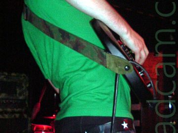 A man with lank dark hair, wearing a bright green t-shirt, standing facing to the right. He is playing a black electric guitar, attached to a green and brown camouflage strap slung over his left shoulder. He is wearing a blue pair of jeans, which are barely held up by a black belt with white stars, his black boxer shorts hanging out over the top of his trousers. Stacks of amplifiers, some with red lights, line the black wall behind him.
