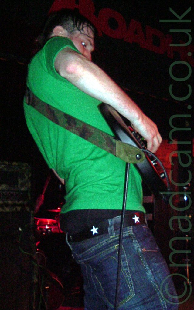A man with lank dark hair, wearing a bright green t-shirt, standing facing to the right. He is playing a black electric guitar, attached to a green and brown camouflage strap slung over his left shoulder. He is wearing a blue pair of jeans, which are barely held up by a black belt with white stars, his black boxer shorts hanging out over the top of his trousers. Stacks of amplifiers, some with red lights, line the black wall behind him.