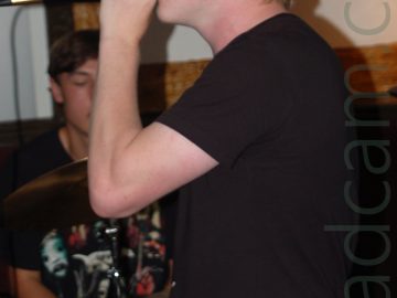 A young man with shaggy blond hair, wearing a black t-shirt and trousers, and holding a microphone to his mouth with his left hand as he faces to the left. Behind him, another young man with short brown hair and wearing a black t-shirt, sits behind a drum-kit, with wood-lined white walls behind him.