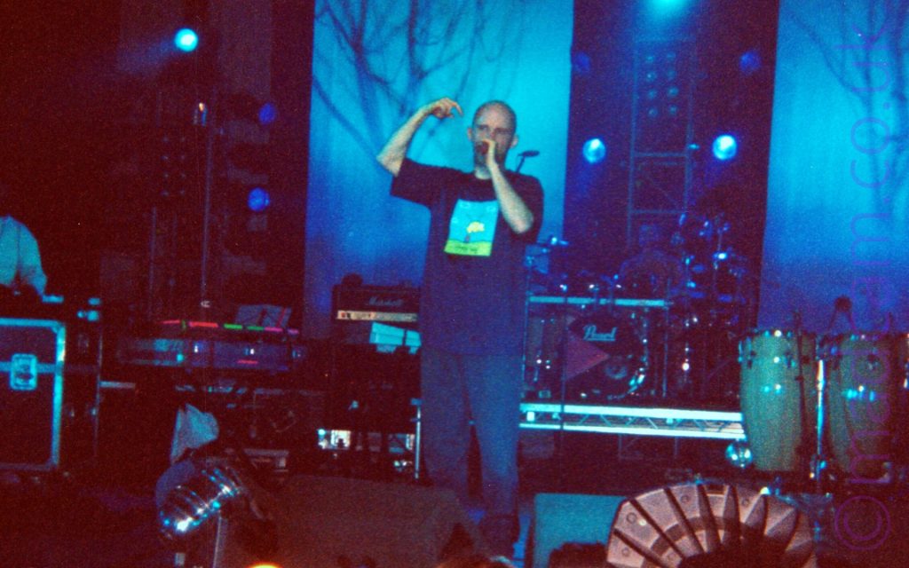 A man standing on stage - he has a shaven head, an is wearing a blue t-shirt with a hand-drawn square image of a cartoon character in the middle of the chest, and a dark blue pair of trousers. Ge is holding a microphone up to his mouth with his left hand, with his right arm in the air by his head. It is a big stage, and although he is surrounded by people, he almost seems alone. A man can be barely discerned sitting behind a black drumkit at the rear of the stage, and another man wearing a white top is almost out of view on the far left of the frame, but other than that, only percussion instruments, racks of amplifiers, and stage lights can be seen.