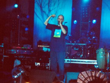 A man standing on stage - he has a shaven head, an is wearing a blue t-shirt with a hand-drawn square image of a cartoon character in the middle of the chest, and a dark blue pair of trousers. Ge is holding a microphone up to his mouth with his left hand, with his right arm in the air by his head. It is a big stage, and although he is surrounded by people, he almost seems alone. A man can be barely discerned sitting behind a black drumkit at the rear of the stage, and another man wearing a white top is almost out of view on the far left of the frame, but other than that, only percussion instruments, racks of amplifiers, and stage lights can be seen.