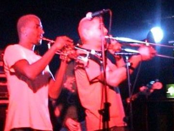 Two men on a stage, one with a shaven head and wearing a white t-shirt, playing a trumpet, and a man with short dark hair, wearing a light-coloured t-shirt and playing a trombone. A microphone on a stand is in front of them. The 2 men are given a red cast by the stage lights, as well as amplifiers in the background, , and the man playing a guitar behind them. Black walls and ceiling fill the frame, with a blue light shining from the other side of the stage.