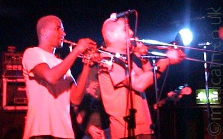 Two men on a stage, one with a shaven head and wearing a white t-shirt, playing a trumpet, and a man with short dark hair, wearing a light-coloured t-shirt and playing a trombone. A microphone on a stand is in front of them. The 2 men are given a red cast by the stage lights, as well as amplifiers in the background, , and the man playing a guitar behind them. Black walls and ceiling fill the frame, with a blue light shining from the other side of the stage.