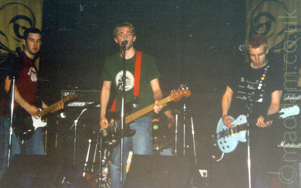4 members of a band performing on a stage. On the left is a man with short dark hair, wearing a mauve t-shirt with white logo and blue sleeves, and a blue pair of jeans, playing a black and white electric guitar. In the middle is a man with short but floppy blond hair and a straggly beard, wearing a green t-shirt with a circular white logo, and blue jeans, playing a black bass guitar. Behind him is a man sitting behind a drum kit - about all that can be seen of him is his left arm, and his blue denim-clad left leg. On the right is a man with short blond hair, arranged into a red spike in the middle of his head, wearing a black t-shirt with lines and seemingly random words, and a dark pair of trousers, playing a powder-blue electric guitar. The 3 men at the front have microphones on stands in front of them. Black walls fill the background, with yellow curtains with circular black logos on either side.