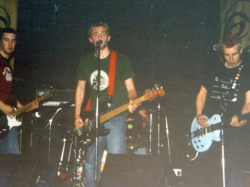 4 members of a band performing on a stage. On the left is a man with short dark hair, wearing a mauve t-shirt with white logo and blue sleeves, and a blue pair of jeans, playing a black and white electric guitar. In the middle is a man with short but floppy blond hair and a straggly beard, wearing a green t-shirt with a circular white logo, and blue jeans, playing a black bass guitar. Behind him is a man sitting behind a drum kit - about all that can be seen of him is his left arm, and his blue denim-clad left leg. On the right is a man with short blond hair, arranged into a red spike in the middle of his head, wearing a black t-shirt with lines and seemingly random words, and a dark pair of trousers, playing a powder-blue electric guitar. The 3 men at the front have microphones on stands in front of them. Black walls fill the background, with yellow curtains with circular black logos on either side.