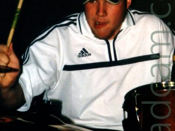 A man wearing a black cap with a red "Ezekiel" logo on the front, wearing a white Adidas top with black pinstripes, sitting behind a red and chrome drumkit. He is holding a wooden drumstick in his right hand, raisesd and ready to strike a cymbal on the left of the frame.