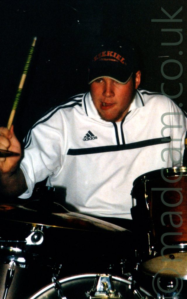 A man wearing a black cap with a red "Ezekiel" logo on the front, wearing a white Adidas top with black pinstripes, sitting behind a red and chrome drumkit. He is holding a wooden drumstick in his right hand, raisesd and ready to strike a cymbal on the left of the frame.