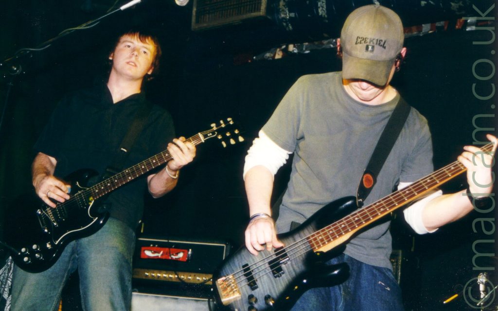 Two men performing on stage as part of a rock band. On the left is a man with short brown hair, wearing a black shirt and a blue pair of jeans, playing a black electric guitar while staring off into the distance. On the right is a man with a green cap on his head, with black text "E2EK1E4" (Ezekiel), a short-sleeved grey t-shirt over a white long-sleeved t-shirt, and a blue pair of jeans, and playing a black bass guitar. Black walls and ceiling fill the rest of the frame.
