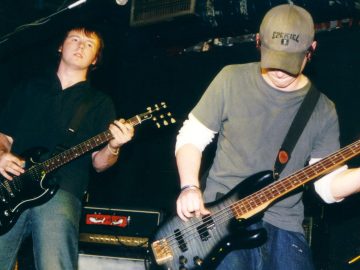 Two men performing on stage as part of a rock band. On the left is a man with short brown hair, wearing a black shirt and a blue pair of jeans, playing a black electric guitar while staring off into the distance. On the right is a man with a green cap on his head, with black text "E2EK1E4" (Ezekiel), a short-sleeved grey t-shirt over a white long-sleeved t-shirt, and a blue pair of jeans, and playing a black bass guitar. Black walls and ceiling fill the rest of the frame.