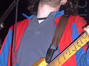 Low-down shot of a man with long, dark, curly hair and a short, dark beard, wearing a red jacket with blue and red sleeves over a grey polo shirt, with a brown guitar strap slung over his shoulder supporting a golden-brown bass guitar. He is singing into a microphone on a stand in front of him. Black walls and ceiling fill the background.
