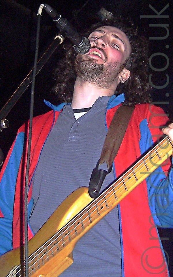 Low-down shot of a man with long, dark, curly hair and a short, dark beard, wearing a red jacket with blue and red sleeves over a grey polo shirt, with a brown guitar strap slung over his shoulder supporting a golden-brown bass guitar. He is singing into a microphone on a stand in front of him. Black walls and ceiling fill the background.