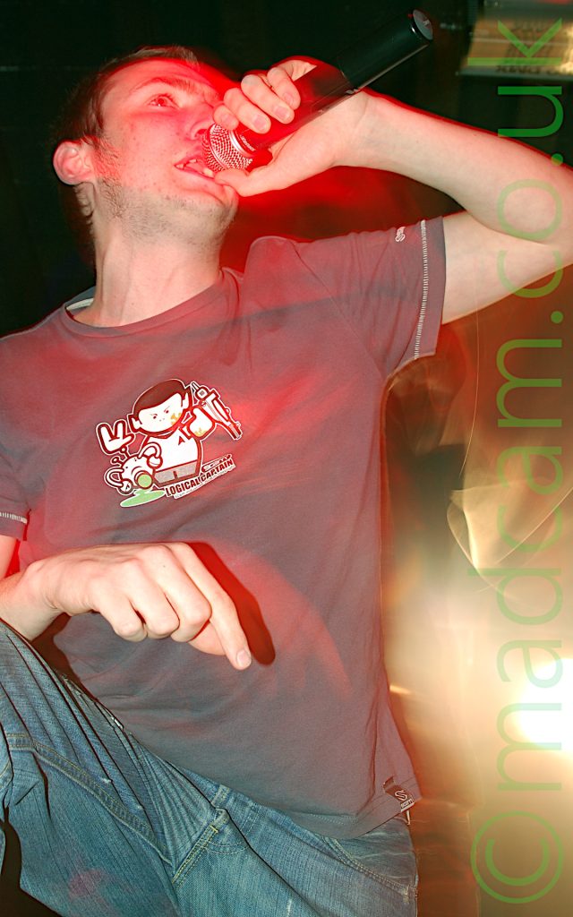 Close-up low shot of a man with short brown hair, wearing a grey with a logo of Spock from Star trek and the words "Logical Captain", as well as a blue pair of jeans. His right leg is leaning on an out-of-shot monitor speaker, with his right arm resting on his knee, while he is singing into a microphone in his left hand while staring off to the right of the frame. A long exposure and his movement leave red streaks where various body parts have moved, with an explosion of green light in the bottom right of the photo. Black ceiling fills the rest of the frame.