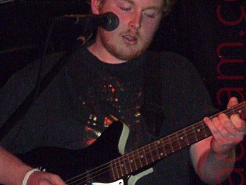 A man with fairly short, slightly curly brown hair and a straggly beard, wearing a black t-shirt with a gold foil logo on the front, playing a black and white guitar with mint green sides., while looking down at his instrument. A microphone on a stand is positioned in front of his face. Black walls and ceiling, with black, vaulting roof beams and white trunking, fill the background.