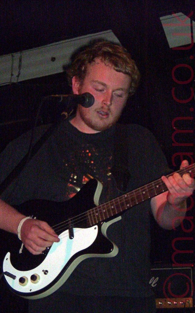 A man with fairly short, slightly curly brown hair and a straggly beard, wearing a black t-shirt with a gold foil logo on the front, playing a black and white guitar with mint green sides., while looking down at his instrument. A microphone on a stand is positioned in front of his face. Black walls and ceiling, with black, vaulting roof beams and white trunking, fill the background.