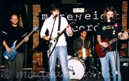 Four members of a band performing on stage. On the left is a man with a shaven head, wearing a black t-shirt and grey trousers, playing a metallic blue bass guitar. Next to him is a man with dark, slightly floppy hair, wearing a white shirt and a blue pair of jeans, playing a black and white electric guitar. At the back is a man with short dark hair, wearing a grey top with a black letter "D" in the middle, sitting behind a gold-coloured drum kit. On the right is a man with long brown hair, wearing a black t-shirt and a blue pair of jeans, playing a red electric guitar. Red brick walls behind them are draped with large, black pieces of cloth, the one behind the drummer has the words "More Weight Records" printed in white, although large parts of this are obscured by the band.