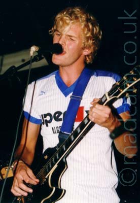 A man with blond hair, wearing a white t-shirt with blue piping on the sleeves and neck, and black pinstripes on the front, with large if indistinguishable black text across the chest. He is playing a black electric guitar, while singing into a mic on a stand in front of his face. Black walls and ceiling fill the background.