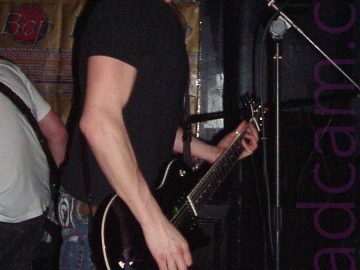 A man with short dark hair standing facing to the right of the frame. He is wearing a black t-shirt and a blue pair of jeans, and playing a black electric guitar with a purple audio cable coming out of the base. A microphone is on a stand in front of him, and he is singing in to it. A person wearing a white t-shirt and black trousers can be seen facing away from the camera on the left of the frame. One of the walls behind is partly covered with brown posters advertising an event called "The Bop". The rest of the walls are black, filling the last of the background.