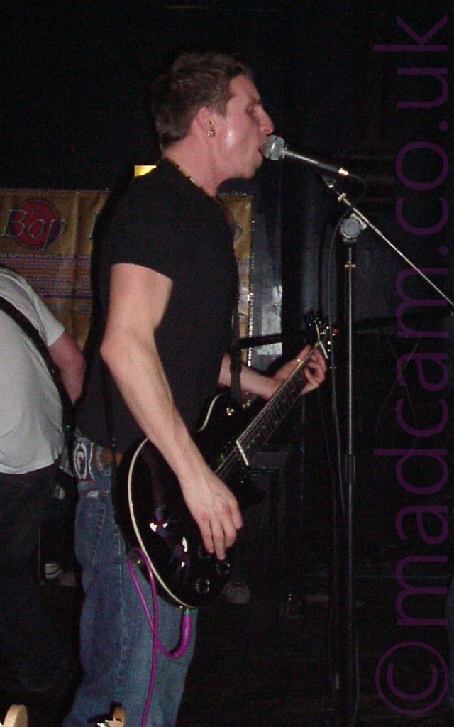 A man with short dark hair standing facing to the right of the frame. He is wearing a black t-shirt and a blue pair of jeans, and playing a black electric guitar with a purple audio cable coming out of the base. A microphone is on a stand in front of him, and he is singing in to it. A person wearing a white t-shirt and black trousers can be seen facing away from the camera on the left of the frame. One of the walls behind is partly covered with brown posters advertising an event called "The Bop". The rest of the walls are black, filling the last of the background.