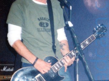 A man with short, slight;y spiky hair, wearing a green t-shirt over a white long-sleeved t-shirt and a blue pair of jeans, playing a black electric guitar and singing in to a microphone on a stand in front of him. A small amplifier stack is on the floor behind him on the left of the frame. Purple walls and black ceiling fill the rest of the frame.