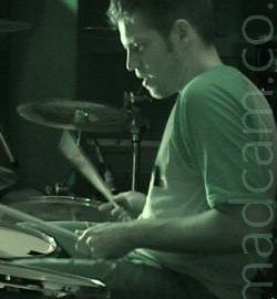 A man sitting behind a drum kit, with a drum stick in each hand that are blurred slightly as they move. He has short, slightly spiky hair, is wearing a light-coloured t-shirt and dark trousers, and is sitting facing to the left. The whole frame has a green tint.